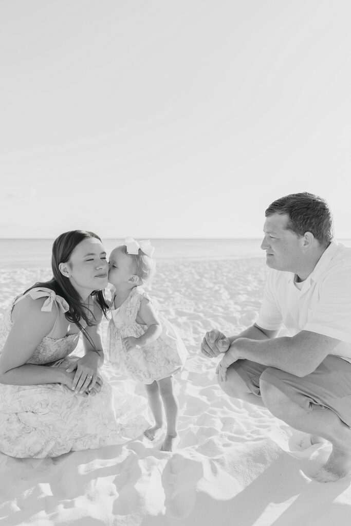 A black and white image of a little girl giving her mom a kiss while her dad watches on Destin beach