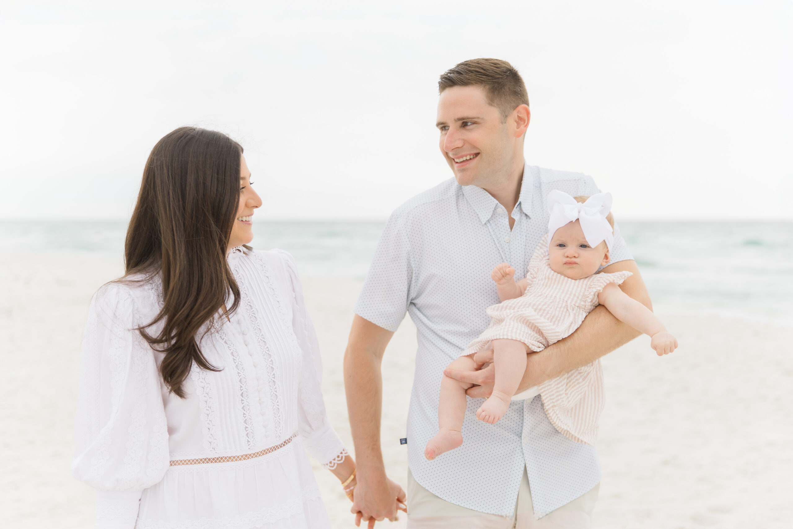 A family of three poses together on a sandy beach during a fall photoshoot. The warm, golden light of the setting sun casts a soft glow, while the family sits on a cozy blanket surrounded by autumn-themed props. The baby, dressed in a snug outfit, smiles as the parents and older sibling look lovingly at one another, capturing the joy and warmth of the moment.